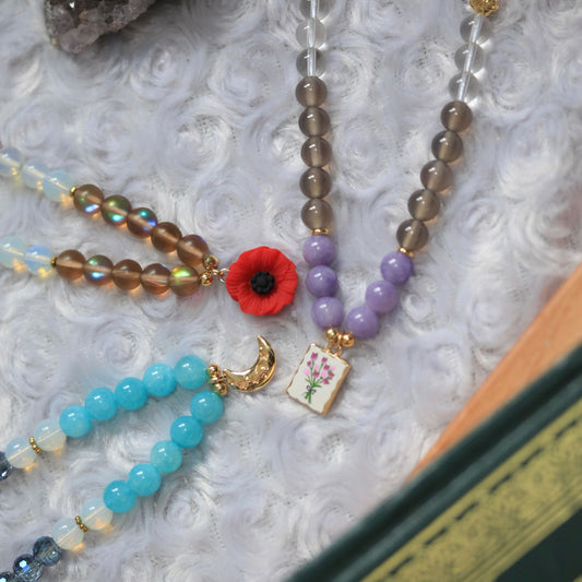 A close-up of several beaded prayer beads arranged on a white textured fabric surface. One prayer beads has translucent smoky brown beads, purple beads, a red poppy flower charm, and a small floral tag.